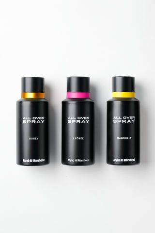 Three black cylindrical perfume spray bottles labeled 'ALL OVER SPRAY' in white text, arranged side by side on a white background. The bottles have black caps and are labeled Honey, Lychee, and Magnolia, with gold, pink, and yellow bands around their necks respectively.