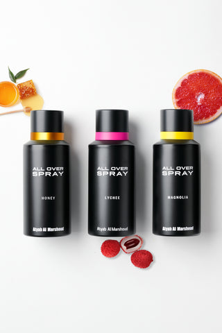 Three black spray bottles labeled 'ALL OVER SPRAY' with scent names Honey, Lychee, and Magnolia, arranged on a white background with fruit props including a grapefruit slice, honey, and raspberries. Brand text 'Atyab Al Marshoud' is visible on the bottles.